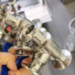 Close-up of a shiny stainless steel sanitary food-grade 3-way ball valve with Tri-Clamp (quick-connect) ends, held by a hand using a blue-handled tool in an industrial workshop.
