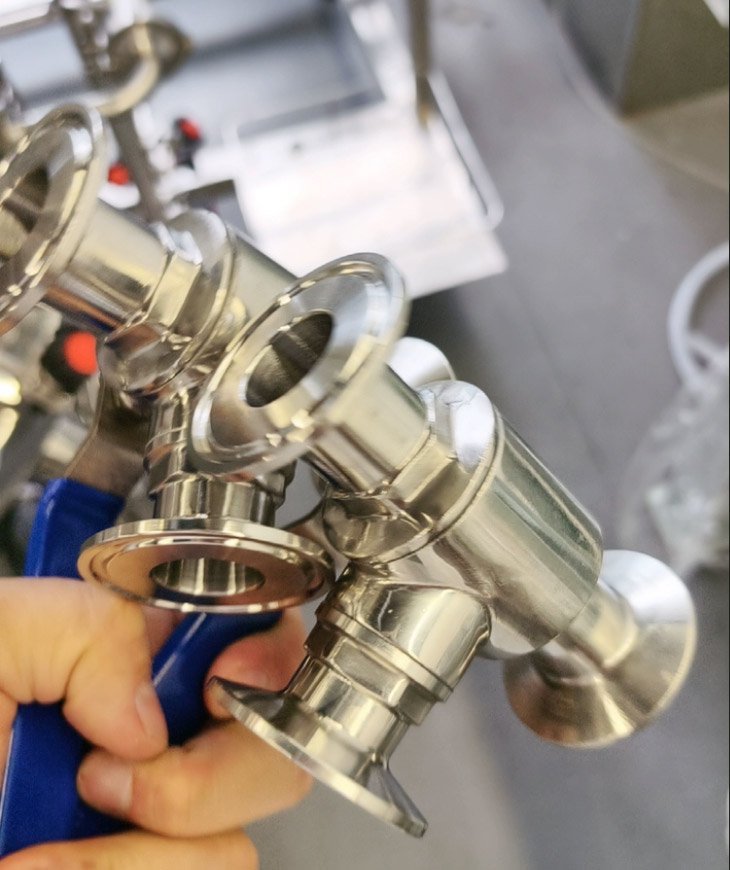 Close-up of a shiny stainless steel sanitary food-grade 3-way ball valve with Tri-Clamp (quick-connect) ends, held by a hand using a blue-handled tool in an industrial workshop.